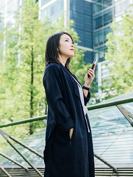 A confident young Asian woman looking at view, holding a smart phone, standing against corporate buildings at the park