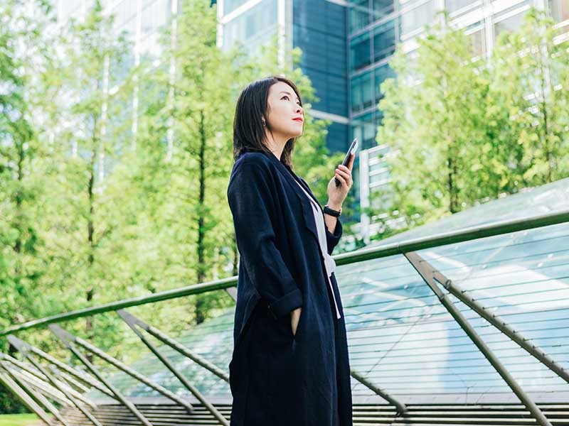 A confident young Asian woman looking at view, holding a smart phone, standing against corporate buildings at the park