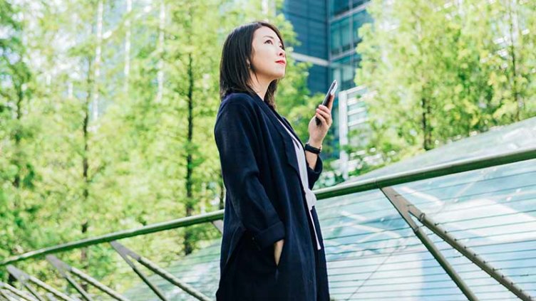 A confident young Asian woman looking at view, holding a smart phone, standing against corporate buildings at the park