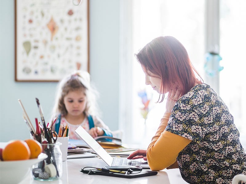 A women working from home on laptop and her daughter sitting nearby and studying